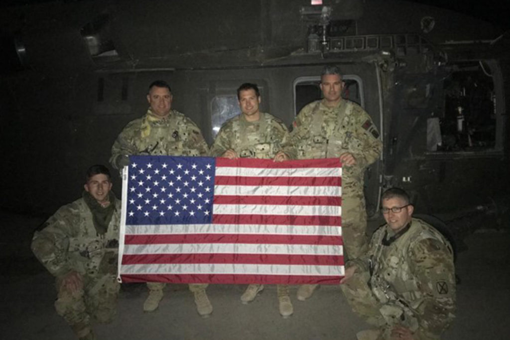 Soldiers from G Company 2-238th General Support Aviation Battalion, as part of Task Force Dragon while deployed to Afghanistan, pose for a photo during a mission with a flag flown to be sent home to their family members, Feb. 6, 2020. The training was done to comply with Army aviation regulations regarding rated and non-rated crewmember requirements. (U.S. Army National Guard courtesy photo)